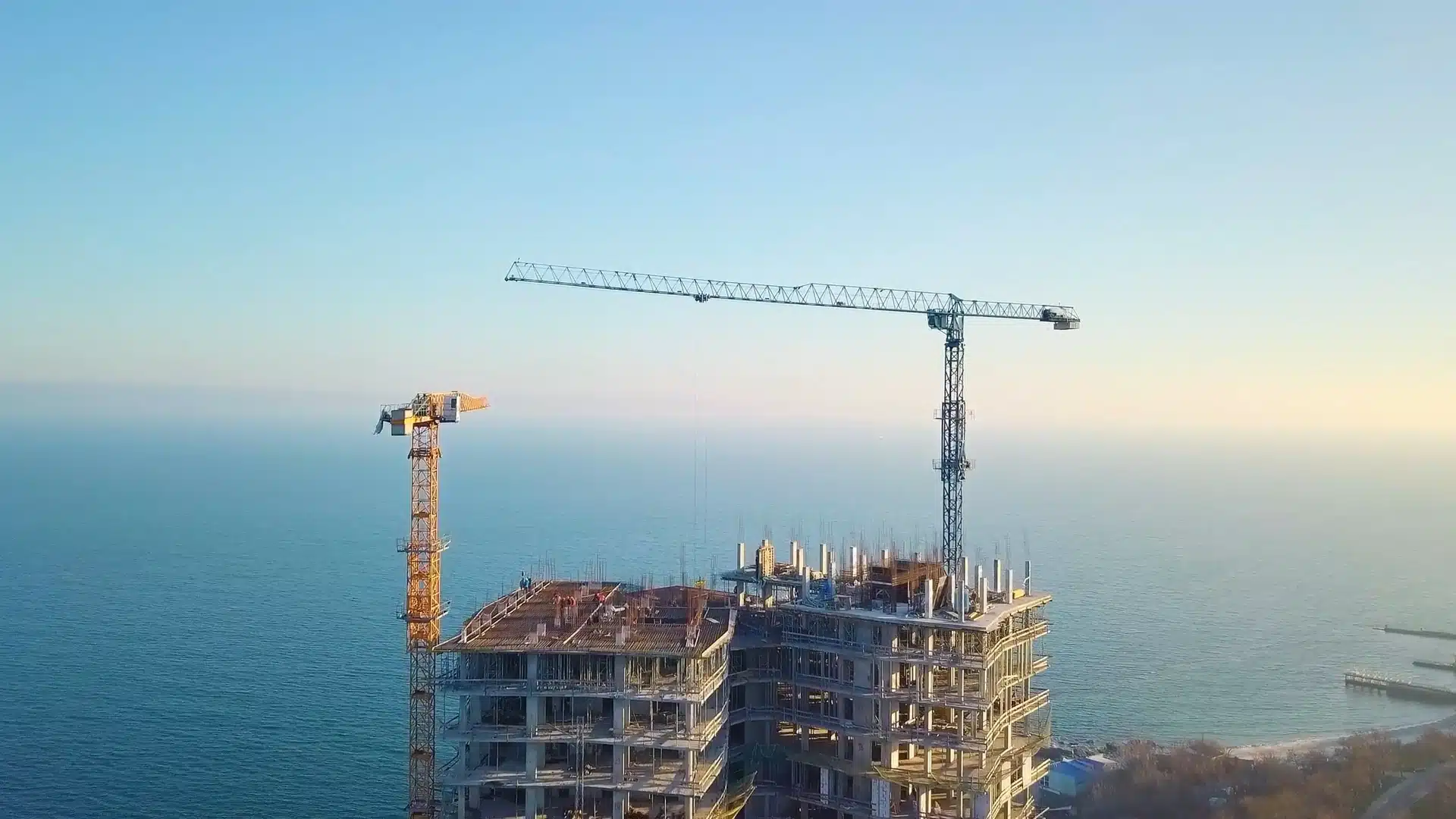 Luftaufnahme einer Baustelle am Meer: Ein mehrstöckiger Rohbau wird errichtet, zwei Turmdrehkrane stehen über dem Gebäude, im Hintergrund sind Meer und klarer Himmel zu sehen.